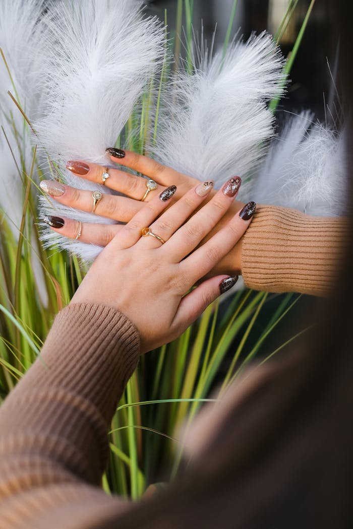 Close-up of a woman's hands adorned with elegant jewelry and stylish nail art against a decorative feather background.